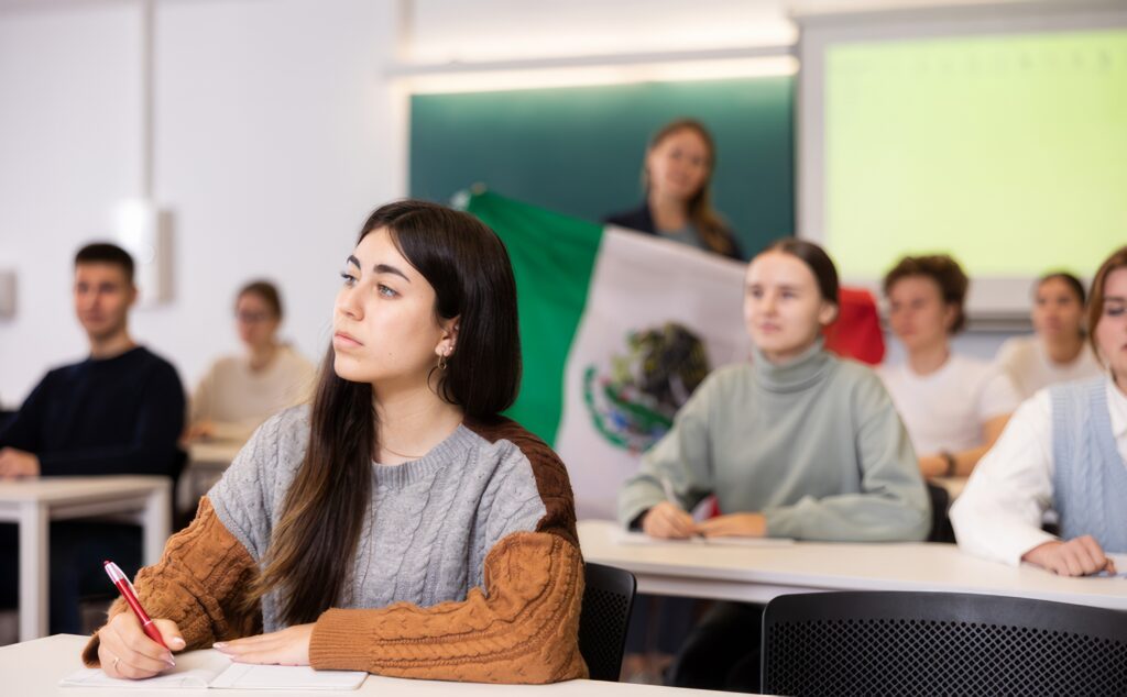 Students sit in classroom, while teacher stands behind them with flag of Mexico